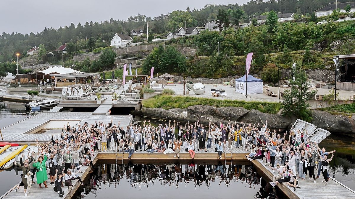 A large group pf people standing on a U-shaped pier, their hands lifted in the air surrounded by water. A tree-covered hill in the background. Photo.
